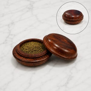 Op de afbeelding: Een ronde, donkerbruine houten container met een gepolijste afwerking, open en gevuld met kleine, gedroogde groene kruiden. Een aparte cirkelvormige afbeelding toont dezelfde houten container met het deksel stevig gesloten.