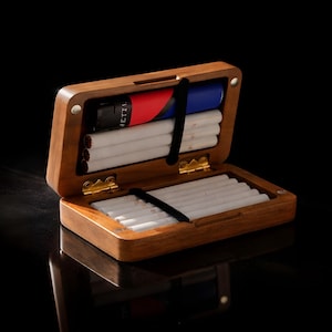 May include: Open wooden cigarette case with a hinged lid, revealing rows of cigarettes and a lighter. The case is made of a warm-toned wood, with gold-colored hinges and a black elastic band to hold the cigarettes in place. The lighter has red, black, and blue accents.