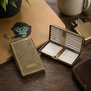 Puede incluir: Una pitillera rectangular de latón con un intrincado grabado floral en el exterior y una placa rectangular en blanco. La pitillera se muestra cerrada y abierta, revelando dos filas de cigarrillos blancos sujetos por una barra metálica.