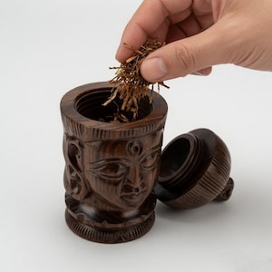 Puede incluir: Un recipiente de madera de color marrón oscuro tallado con forma de rostro y una tapa a juego. El recipiente presenta intrincados patrones de veta de madera y un interior hueco. Se está colocando una pequeña cantidad de tabaco marrón suelto.