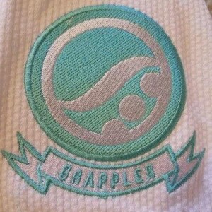 May include: Embroidered patch with a teal and silver design on a white textured fabric. The patch features a circular emblem with a wave-like pattern and the word "GRAPPLER" on a banner below. The design is outlined in teal.