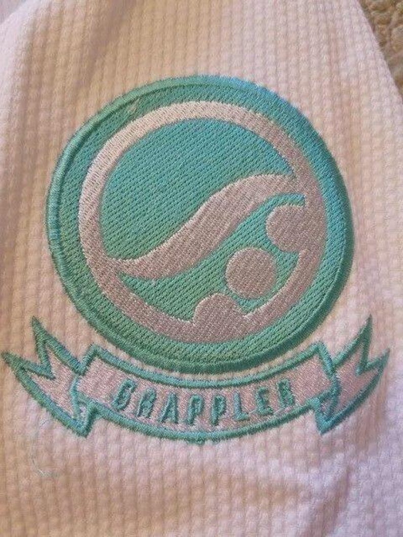May include: Embroidered patch with a teal and silver design on a white textured fabric. The patch features a circular emblem with a wave-like pattern and the word "GRAPPLER" on a banner below. The design is outlined in teal.