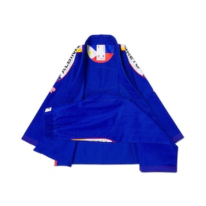 May include: A royal blue Brazilian Jiu-Jitsu gi featuring white and red accents on the sleeves and collar. The interior neck label displays the letter S, and the inner collar lining features a circular graphic with a yellow sun and red and blue segments.
