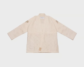May include: Off-white martial arts gi with a v-neck collar and long sleeves. The gi features embroidered patterns on the sleeves and lower front. The brand name is visible on the collar and lower front. The gi is laid flat on a light background.