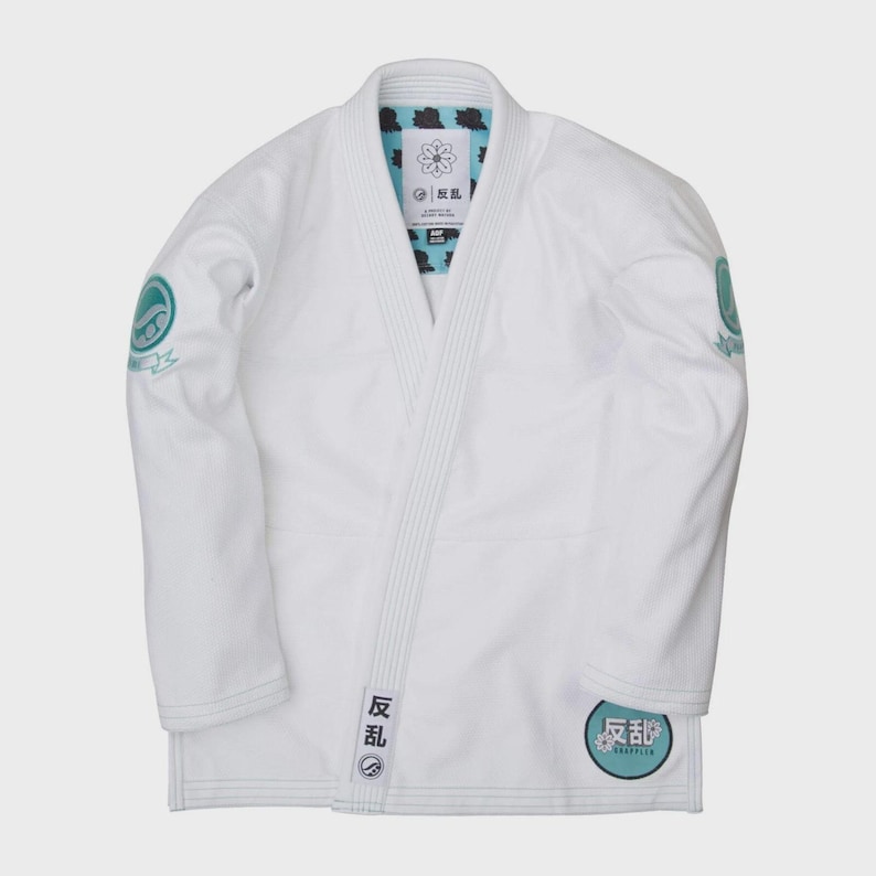 May include: A white martial arts gi with teal and black accents. The gi features patches with Japanese text and circular logos on the sleeves and lower front. The gi is laid flat, showcasing its design and construction.