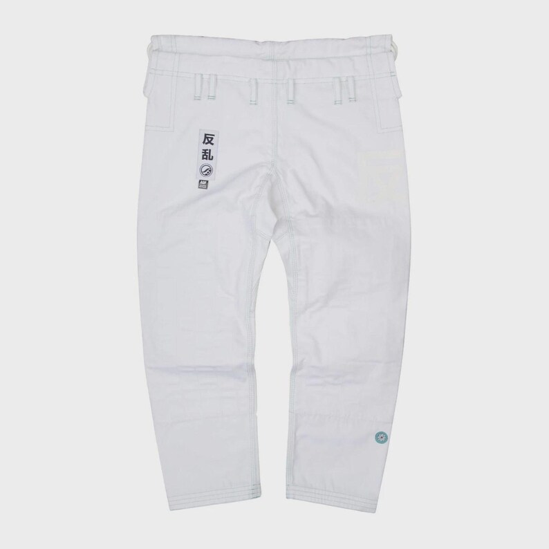 May include: White martial arts pants with visible stitching and a drawstring waist. The pants feature a small patch with Japanese text on the left leg and a circular emblem on the right leg. The pants are made of a durable material.