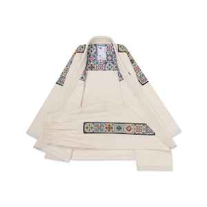 Puede incluir: Un gi de artes marciales color blanco hueso con un patrón de azulejos multicolor en los hombros y una franja horizontal a juego en el pantalón. El cuello interior muestra una etiqueta con el texto ML. La tela tiene costuras reforzadas visibles.