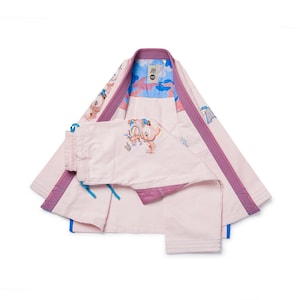 Puede incluir: Un uniforme de artes marciales rosa claro con ribetes morados y detalles bordados. El uniforme incluye una chaqueta y un pantalón, con un interior azul y estampado. La chaqueta tiene cuello y mangas, y el pantalón tiene un cordón.