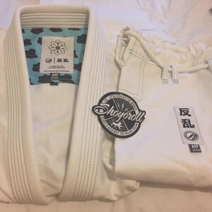 May include: White Jiu-Jitsu uniform set, including a jacket and pants. The jacket has a light blue patterned lining and a patch with Japanese characters. A circular patch with the text "Shoyoroll" is also visible.