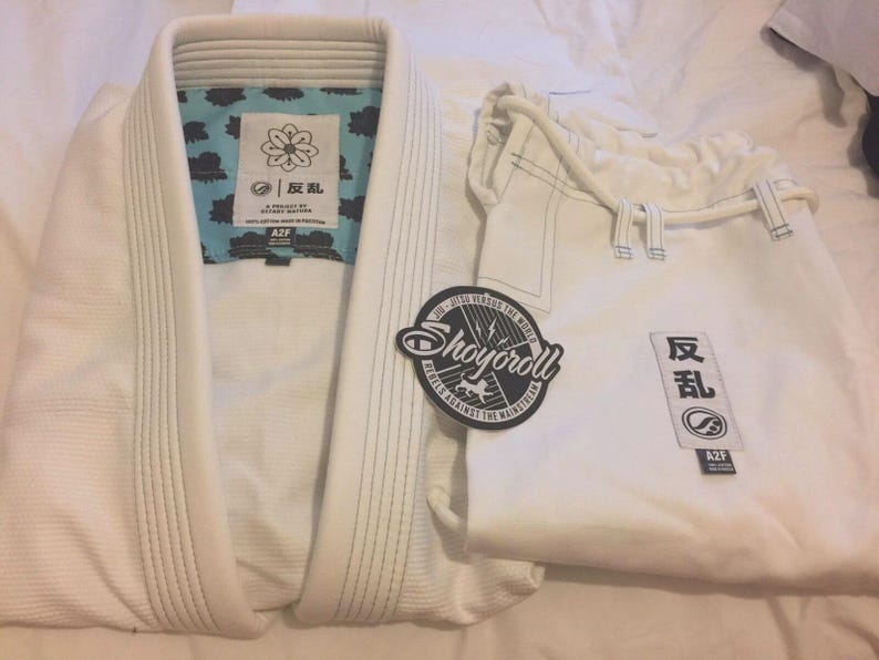 May include: White Jiu-Jitsu uniform set, including a jacket and pants. The jacket has a light blue patterned lining and a patch with Japanese characters. A circular patch with the text "Shoyoroll" is also visible.