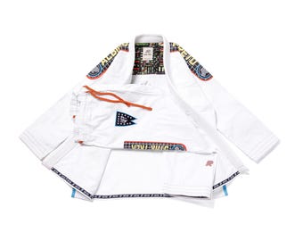 Puede incluir: Un uniforme de artes marciales blanco de manga larga con pantalones a juego y cordón naranja. La prenda incluye parches coloridos con patrones de cuadrícula, el texto STAR TREK y un emblema UFP en los pantalones y en las áreas de los hombros.
