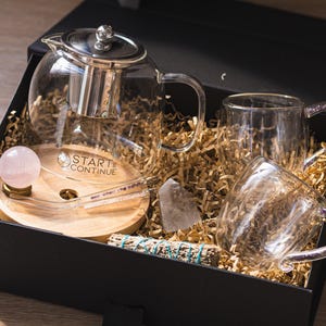 May include: A black gift box filled with a glass teapot, two glass mugs, a wooden tray with a rose quartz sphere, a crystal, and a bundle of sage. The box is lined with gold-colored paper.