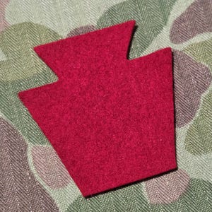 May include: A red felt keystone shape, a symbol of Pennsylvania, is displayed against a camouflage background. The keystone is a solid, vibrant red, with a textured surface. The background features a green, brown, and gray camouflage pattern.