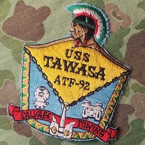 May include: A yellow, blue, and red embroidered patch with the text "USS TAWASA ATF-92" and the words "SAVAGE TOWING" on a red banner. The patch features a Native American figure wearing a headdress.