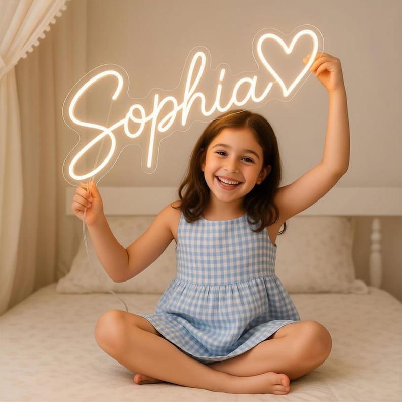 Puede incluir: Una ni&ntilde;a sonriente sostiene un letrero de ne&oacute;n con la palabra "Sophia" y un coraz&oacute;n. El letrero emite una luz blanca c&aacute;lida. La ni&ntilde;a lleva un vestido a cuadros azul y blanco y est&aacute; sentada en una cama.