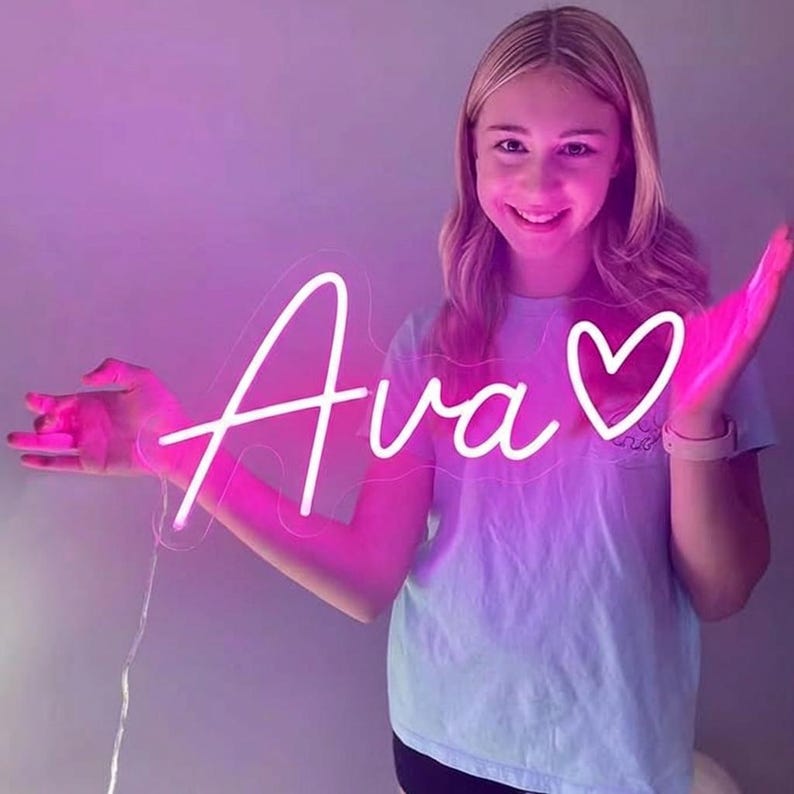 Puede incluir: Un letrero de ne&oacute;n blanco con el nombre "Ava" y un coraz&oacute;n. La persona sostiene el letrero frente a ella, creando un brillo rosado. El fondo es un degradado de rosa y morado. Ideal para un dormitorio o evento.