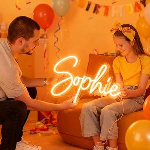 Puede incluir: Un letrero de ne&oacute;n con el nombre "Sophie" en cursiva. El letrero es sostenido por una persona y una ni&ntilde;a. El fondo incluye globos y una pancarta de "Feliz Cumplea&ntilde;os", lo que sugiere una celebraci&oacute;n de cumplea&ntilde;os.