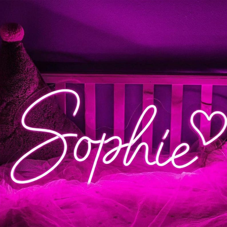 Puede incluir: Un letrero de ne&oacute;n blanco con el nombre "Sophie" en cursiva y un coraz&oacute;n al final. El letrero brilla sobre un fondo morado oscuro. Ideal para una guarder&iacute;a o dormitorio.
