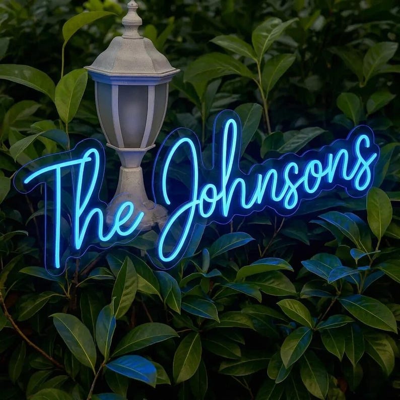 May include: A blue neon sign with the text "The Johnsons" in a cursive font. The sign is surrounded by green foliage and is illuminated by a vintage-style outdoor lamp.