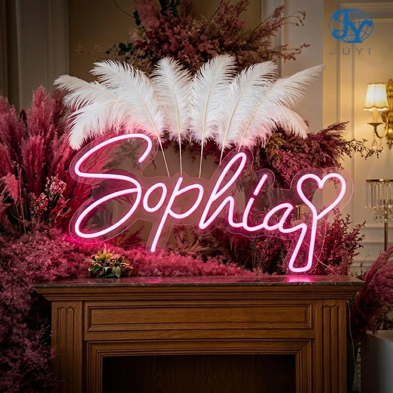May include: A neon sign spelling "Sophia" in a cursive font, with a heart replacing the "a". The sign is lit with a bright pink light. White feathers and pink floral arrangements are in the background, with a dark brown wooden mantelpiece in the foreground.