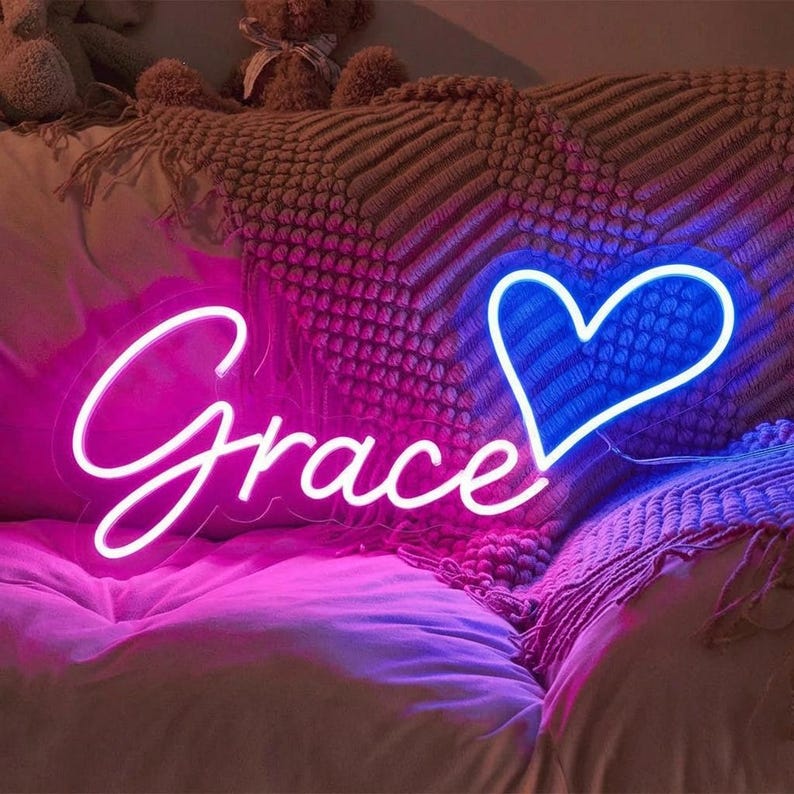 Puede incluir: Un letrero de ne&oacute;n con el nombre "Grace" en fuente cursiva, iluminado en rosa brillante. Una forma de coraz&oacute;n azul est&aacute; al lado del nombre. El letrero est&aacute; colocado sobre una cama con un degradado rosa y morado.