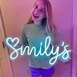 May include: A neon sign in a cursive font that reads "Smily's" in bright blue. The sign has a heart shape at the beginning of the name. The sign is held by a person wearing a green shirt and purple shorts.