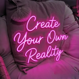 May include: A bright pink neon sign with the words "Create Your Own Reality" in a cursive font. The sign is placed in front of a gray pillow with a decorative tassel. The overall aesthetic is modern and inspirational.