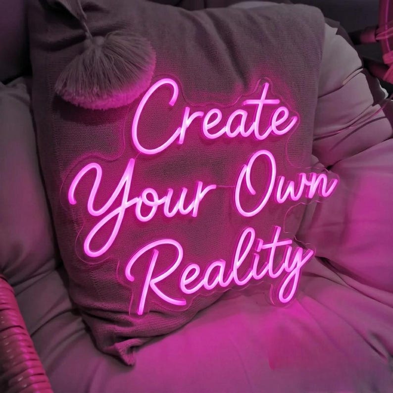 May include: A bright pink neon sign with the words "Create Your Own Reality" in a cursive font. The sign is placed in front of a gray pillow with a decorative tassel. The overall aesthetic is modern and inspirational.