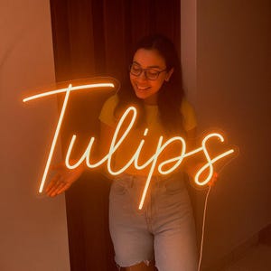 May include: A neon sign in a cursive font that spells "Tulips" in a warm, glowing orange color. The sign is held up in front of a person. The background is a neutral brown.