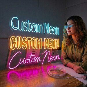 May include: Three neon signs spelling "Custom Neon" in blue, yellow, and pink. The signs are lit up and placed on a wooden surface. A woman wearing glasses is looking at the signs.