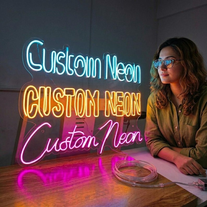 May include: Three neon signs spelling "Custom Neon" in blue, yellow, and pink. The signs are lit up and placed on a wooden surface. A woman wearing glasses is looking at the signs.
