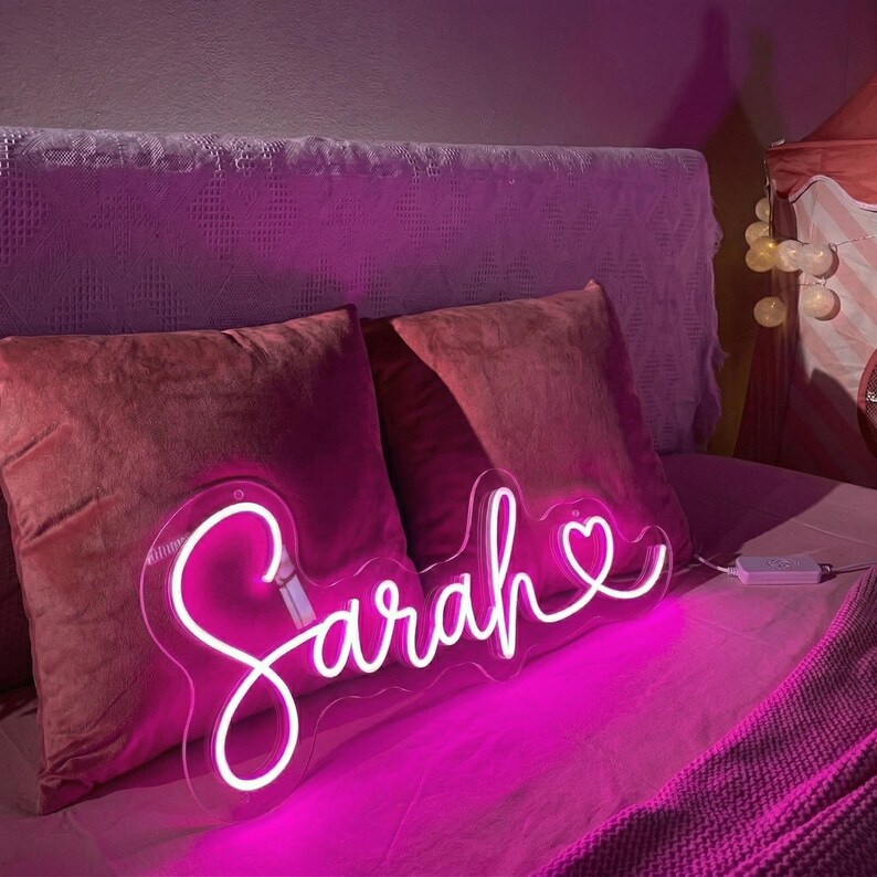 Puede incluir: Un letrero de ne&oacute;n con el nombre "Sarah" en cursiva, con un s&iacute;mbolo de coraz&oacute;n. El letrero emite un brillo rosa brillante y est&aacute; colocado en una cama con almohadas y ropa de cama rosas. El letrero se alimenta con una caja de control blanca.