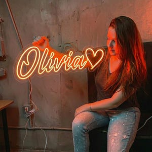 Puede incluir: Un letrero de ne&oacute;n en luz naranja c&aacute;lida muestra el nombre "Olivia" en escritura cursiva, seguido de un s&iacute;mbolo de coraz&oacute;n. El letrero lo sostiene una persona en una habitaci&oacute;n con poca luz y una pared de hormig&oacute;n.