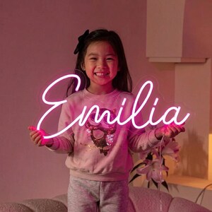 Puede incluir: Un letrero de ne&oacute;n en fuente cursiva que deletrea "Emilia" en rosa brillante. El letrero lo sostiene un ni&ntilde;o sonriente que lleva una sudadera rosa y un pantal&oacute;n de ch&aacute;ndal gris. El fondo es rosa suave.