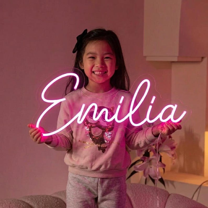 Puede incluir: Un letrero de ne&oacute;n en fuente cursiva que deletrea "Emilia" en rosa brillante. El letrero lo sostiene un ni&ntilde;o sonriente que lleva una sudadera rosa y un pantal&oacute;n de ch&aacute;ndal gris. El fondo es rosa suave.