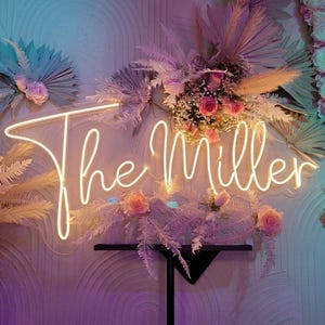 May include: A neon sign that reads "The Miller" in a cursive font, surrounded by floral arrangements and decorative foliage. The sign is illuminated in a warm white color, with pink roses and dried plants.