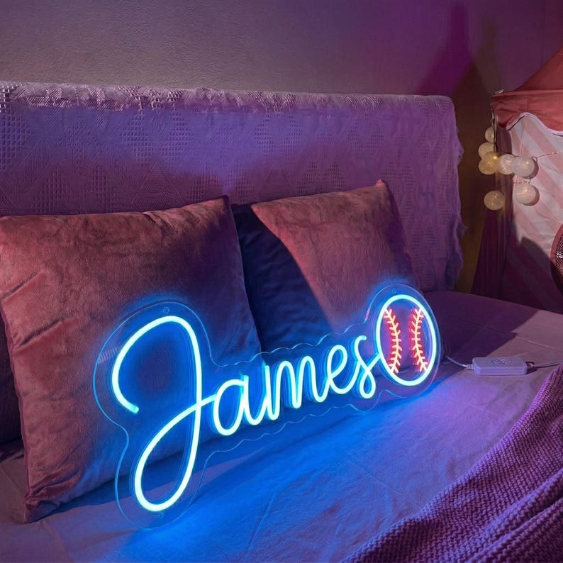 Puede incluir: Un letrero de ne&oacute;n azul con el nombre "James" en cursiva, con un gr&aacute;fico de b&eacute;isbol. El letrero est&aacute; sobre un respaldo transparente y se coloca sobre una cama con almohadas granates y una colcha morada. El gr&aacute;fico de b&eacute;isbol es rojo y blanco.