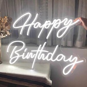 May include: A white neon sign that reads "Happy Birthday" in a cursive font. The sign is held by a hand and is against a window with white curtains. The sign is a decorative item for a birthday celebration.