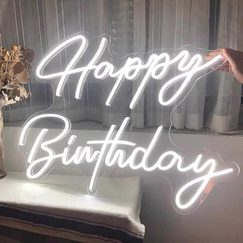 May include: A white neon sign that reads "Happy Birthday" in a cursive font. The sign is held by a hand and is against a window with white curtains. The sign is a decorative item for a birthday celebration.