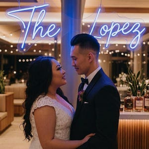 May include: A couple gazing at each other, with a neon sign above them that reads "The Lopez". The woman wears a white lace wedding dress, and the man is in a black tuxedo with a bow tie. The background includes a bar with bottles.