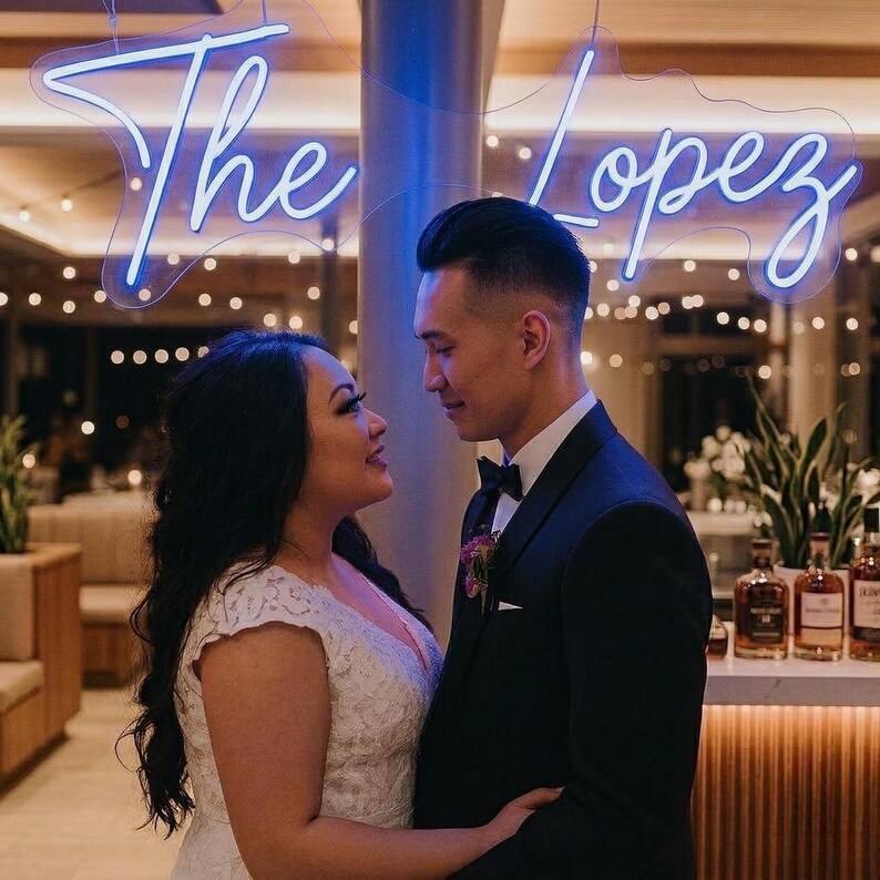 May include: A couple gazing at each other, with a neon sign above them that reads "The Lopez". The woman wears a white lace wedding dress, and the man is in a black tuxedo with a bow tie. The background includes a bar with bottles.