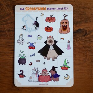 Peut inclure: Une planche d'autocollants avec des illustrations sur le thème d'Halloween. La planche comprend des images de citrouilles, de chats, de capybaras en costumes, de flacons de potion et d'un chaudron. Le texte "the SPOOKYBARA sticker sheet (2)" est en haut.