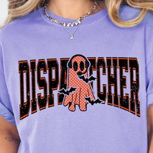 May include: A light purple short-sleeve t-shirt featuring the word DISPATCHER in large black and orange block letters. An orange ghost wearing a headset is positioned in the center of the text, surrounded by small black flying bat silhouettes.