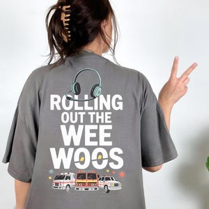 Puede incluir: Camiseta gris con la frase "Rolling Out The Wee Woos" y un diseño de auriculares, una ambulancia, un camión de bomberos y un coche de policía. También se ve el logotipo de Lake Martin Shirt Shack.