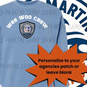 Op de afbeelding: Een lichtblauw sweatshirt met de tekst "wee woo crew" boven een schildvormig embleem. Het embleem luidt "Communications Division" en "Alexander City Police Department". Een oranje ster zegt "Personaliseer naar uw agentschappen patch of laat leeg".