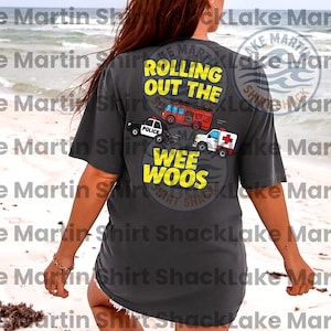 May include: Dark gray t-shirt with a graphic on the back featuring a police car, fire truck, and ambulance. The text reads "Rolling Out The Wee Woos" in yellow. The shirt is a casual style.