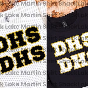 May include: Two t-shirts, one white with black pinstripes and the other black, both featuring the letters "DHS" in a bold, layered design with black and gold colors. The shirts are likely for a school or team.