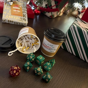 May include: A Christmas-themed image featuring a brown coffee cup with the label "A cup of Xmas Cheer", dice, and wrapped gifts. A second cup is filled with white and gold confetti. Festive and celebratory.