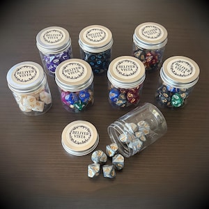 May include: Seven small glass jars filled with colourful polyhedral dice, each with a silver lid and a label that reads "BELIYER VISTA". One jar is tipped over, spilling dice. The dice are various colours, including purple, blue, red, and green.