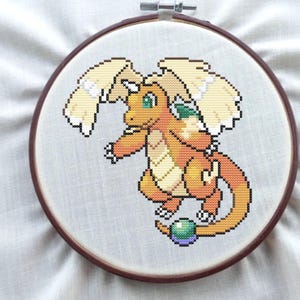 May include: A cross-stitch embroidery of a cartoon dragon in a wooden hoop. The dragon is primarily orange with yellow wings and green eyes. The embroidery is on a light gray fabric.